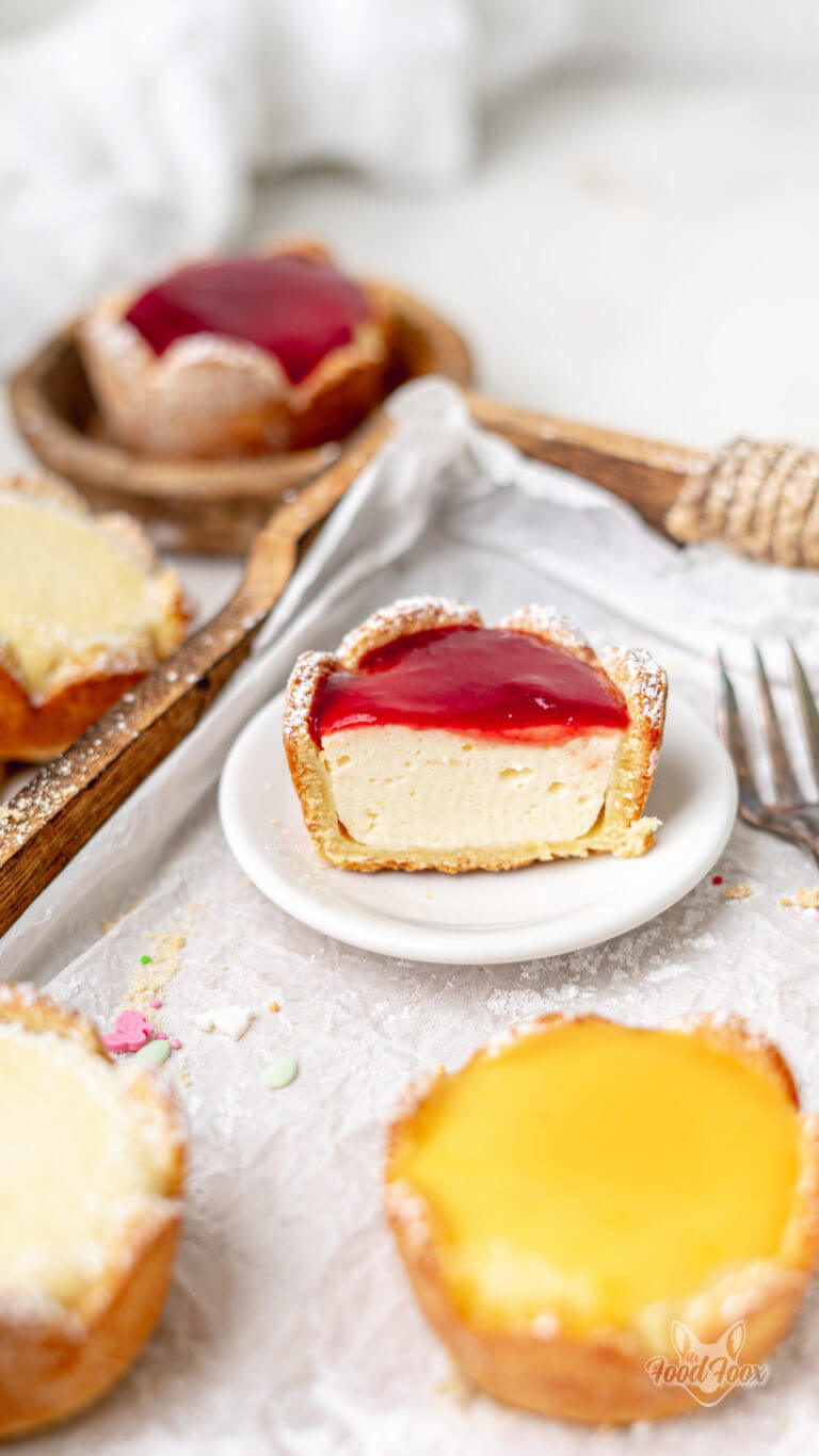 Cheesecake Muffin mit Erdbeerspiegel, angeschnitten, auf einem weißen Teller. Nahaufnahme. Im Vordergrund sind weitere Käsekuchenmuffins zu erkennen.