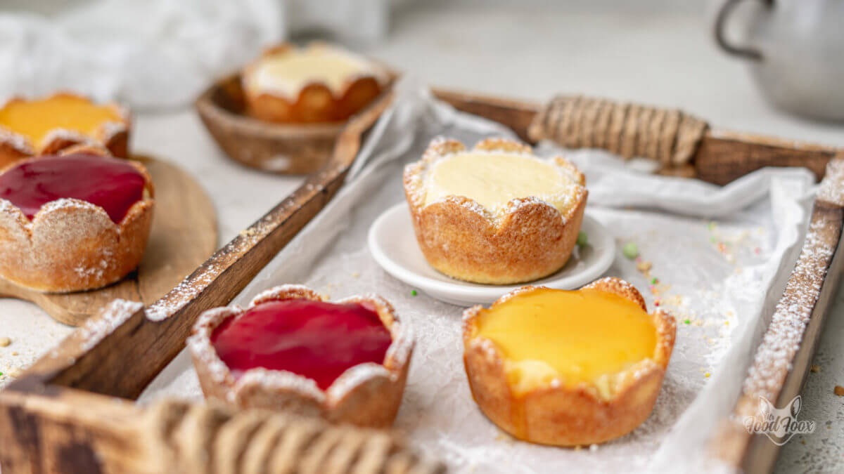 Käsekuchen Muffins mit Mürbeteigboden, Erdbeerspiegel und Puderzucker auf einem Holztablett.