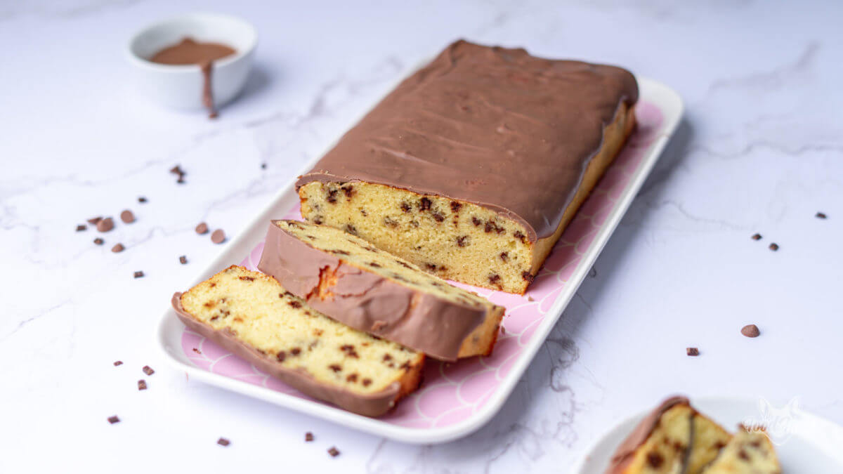 Protein Kuchen mit Schokosplittern und Schokoladenglasur auf einem rosa Servierteller. Der Kuchen wurde angeschnitten und sieht sehr fluffig aus. Im Hintergrund ist ein Schälchen mit Schokolade zu erkennen.