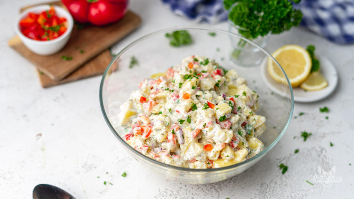 Karlorienarmer Kartoffelsalat mit einem leichten Joghurtdressing, Paprika und Gurke. Anerichtet in einer Glasschüssel. Im Hintergrund ist frische Petersilie, eine Zitrone und Paprika-Würfel erkennbar.