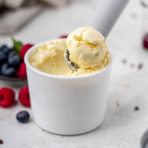 Selbst gemachtes, kalorienarmes Eis in einem weißen Eisbecher. Der EIslöffel steckt drin. Im Hintergrund sind Himbeeren und Blaubeeren erkennbar.