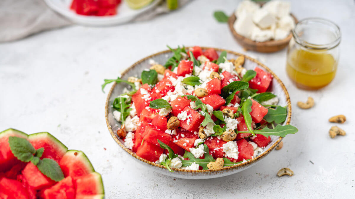 Melone-Feta-Salat mit Minze und Rucola sowie Cashenüssen in eine tiefen Bowl. Ein paar Nüsse liegen auf dem Tisch. Im Hintergrund ist das fruchtige Dressig erkennbar.