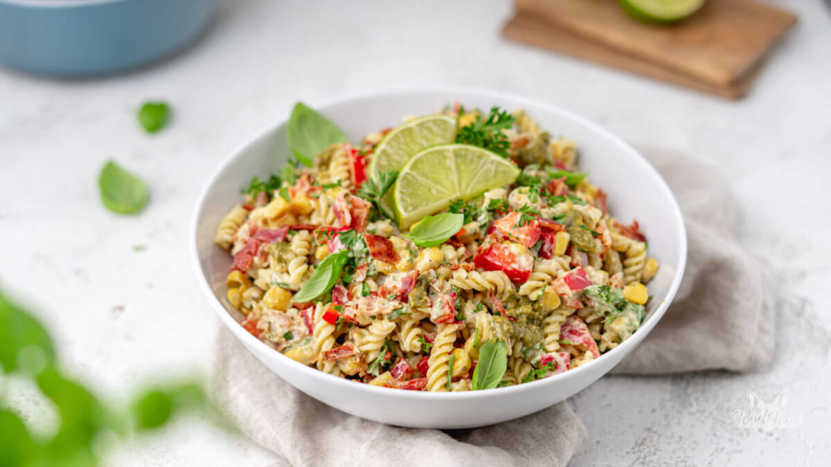 Sommerlicher Nudelsalat mit Joghurt, Paprika, Mais und frischem Basilikum. Angerichtet in einem tiefen Teller, dieser steht auf einem beigen Tuch.