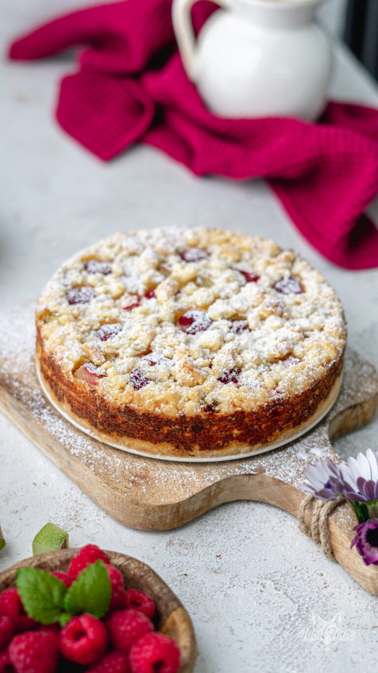 Cremiger Rhabarber-Cheesecake mit zuckerfreien Streuseln. Der Kuchen wurde mit Puderzucker bestreut und steht auf einem hellen Tisch. Im Vordergrund sind frische Himbeeren zu sehen. Im Hintergrund ein rosanes Tuch.