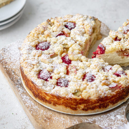 Cremiger Rhabarber-Käsekuchen mit Streuseln. Der Kuchen wurde angeschnitten ein, Stück wird gerade entnommen. Er steht auf einem Holzbrettchen. Hier und da liegt Puderzucker.