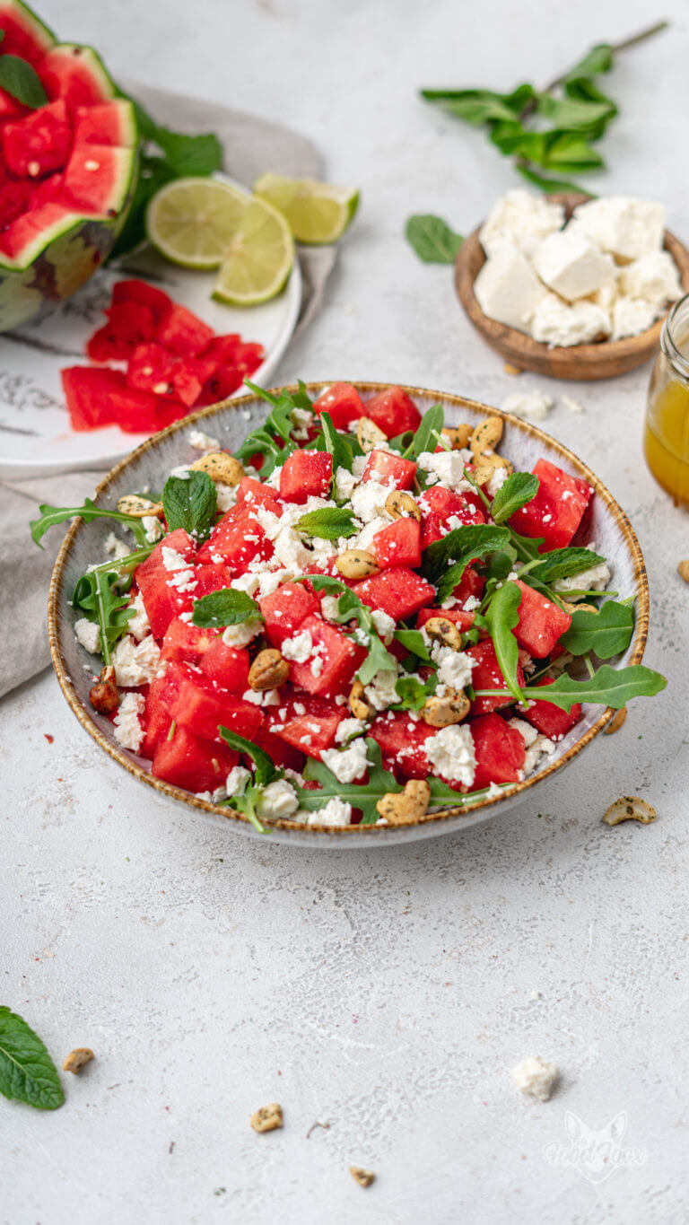 Melonen-Feta-Salat mit Minze und Rucola in einem großen tiefen Teller. Im Hintergrund sind frische Limetten, das fruchtige Dressing und Feta zu sehen.