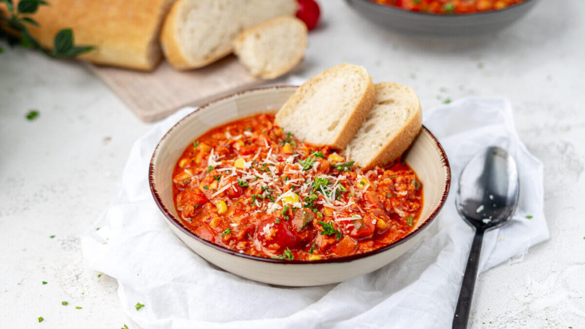 Low Carb Pizzasuppe mit Hackfleisch und viel Gemüse angerichtet in einem tiefen Teller. Zwei Scheiben Low Carb Brötchen liegen an Beilage drin.