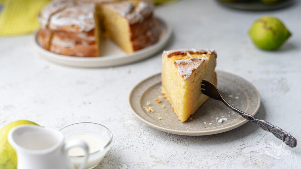 saftiger, kalorienarmer Zitronenkuchen ohne Zucker. Ein Stück steht auf einem Teller. In die Seite ist eine Kuchengabel gepiekt. Im Hintergrund ist der restliche Kuchen sichtbar.