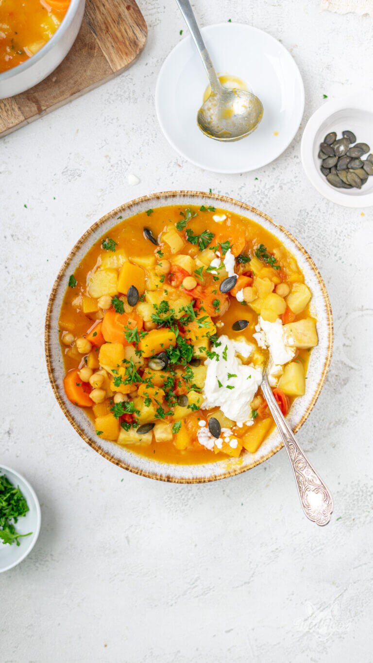herbstlich bunter Kürbis-Kartoffel-Eintopf in einem tiefen Suppenteller garniert mit frischer Petersilie und Saurer Sahne sowie Kübriskernen
