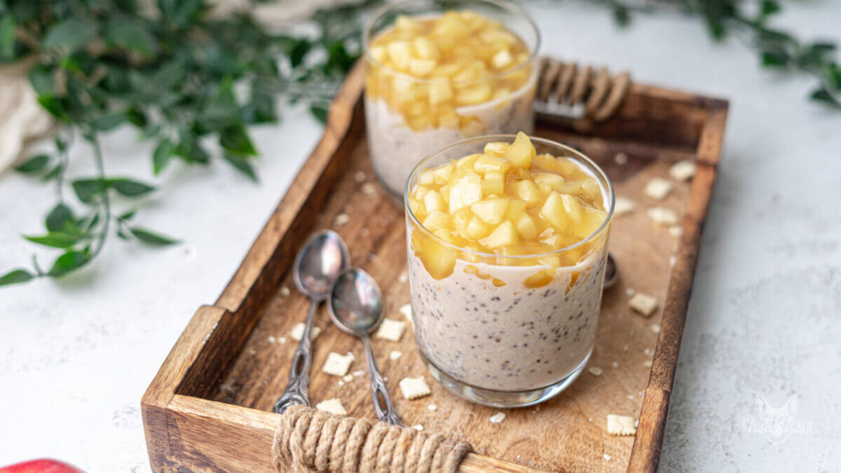 High Protein Obvernight Oats mit Apfel und Zimt in zwei Gläsern. Die Chiassamen bilden ein schönes Muster. Die Gläser mit overnightoats stehen auf einem Holztablatt. Daneben liegen zwei Löffel.