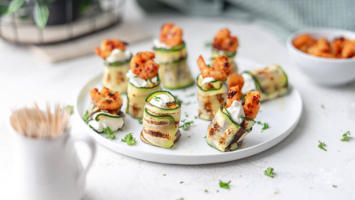 Zucchini-Frischkäse Röllchen auf einem weißen Teller. teilweise mit einer gebratenen Garnele garniert.