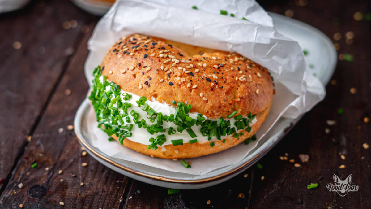 Selbst gemachte high Protein Bagels mit Sesam und Frischkäse in einer weißen servierte auf einem Teller. Im Hintergrund ist ein weiterer Bagel erkennbar.