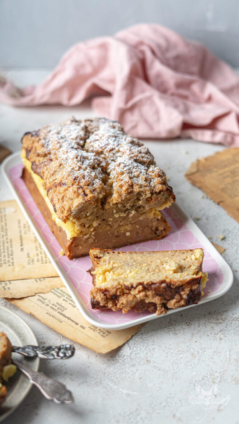 Einfaches proteinreiches Bananenbrot mit Streuseln. Angeschnitten auf einem länglichen Kuchenteller. Im Vordergrund liegt ein Stück.