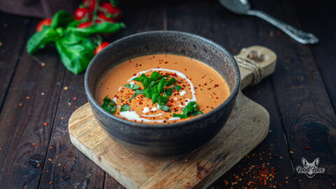 Kalorienarme Tomatensuppe zum Abnehmen in einer tiefen Schale, welche auf einem Holzbrettchen steht. Die Suppe wurde mit Basilikum, frischen Tomatenflocjen und Sahne verfeinert.