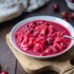 Kirschsuppe mit Klüten auf einem Holzbrett, seitlich aufgenommen. In der weißen Schale schwimmen Kirschen und Mehlklößchen im roten Kirschsaft; frische Kirschen liegen auf dem dunklen Holztisch, ein helles Tuch sorgt für eine weiche Stimmung.